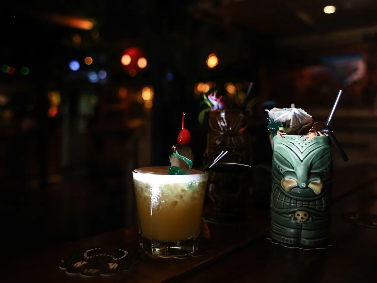 A Mai Tai style cocktail and tiki mugs on a dark bar counter