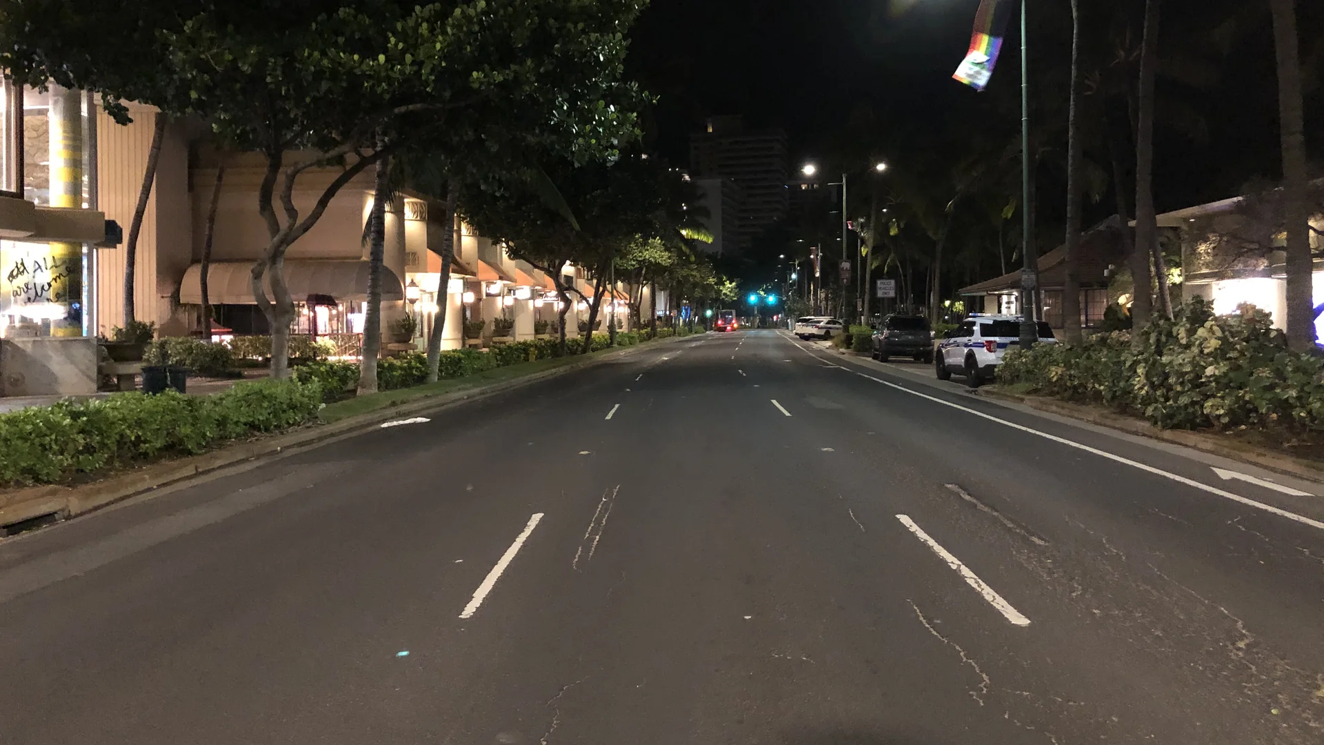 Kalākaua Avenue in Waikiki at night, empty of traffic, palm trees and lit storefronts lining both sides of the street