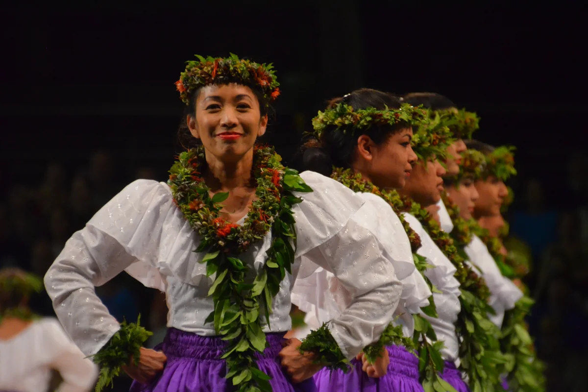 A row of hula dancers in white blouses, purple skirts and flower leis, smiling on a dark stage